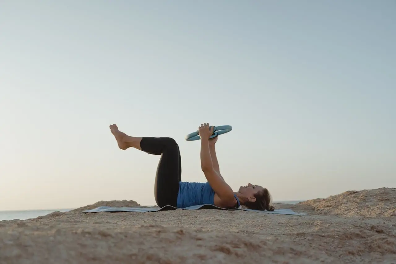 Woman lying on yoga mat, exercising with a ring on a rocky shoreline during daylight.