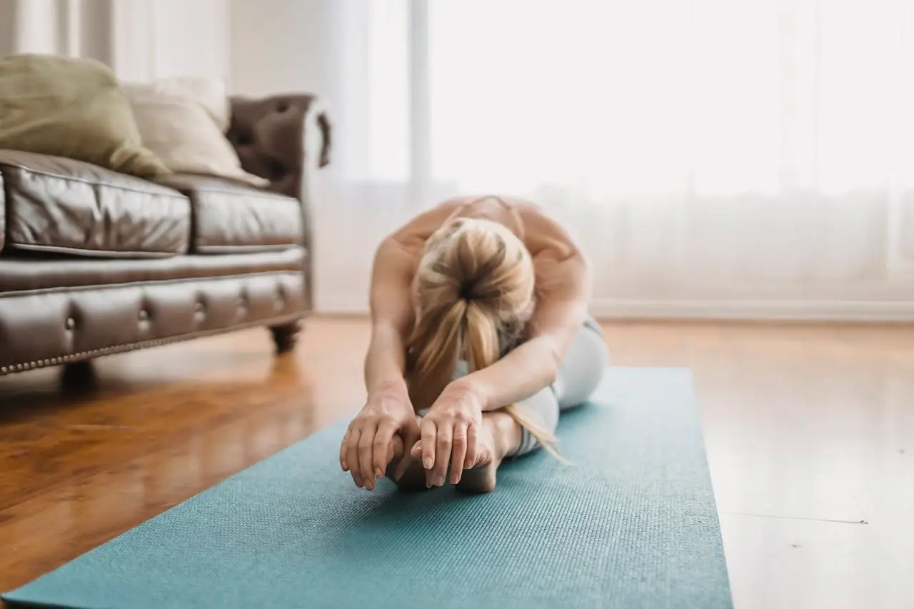 contact-img A woman performs a yoga stretch on a mat in a cozy living room, embracing wellness and relaxation.