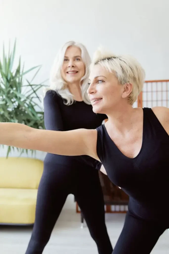 pexels-photo-8374568-8374568 Two senior women practicing yoga indoors, dressed in black, smiling and stretching for fitness.